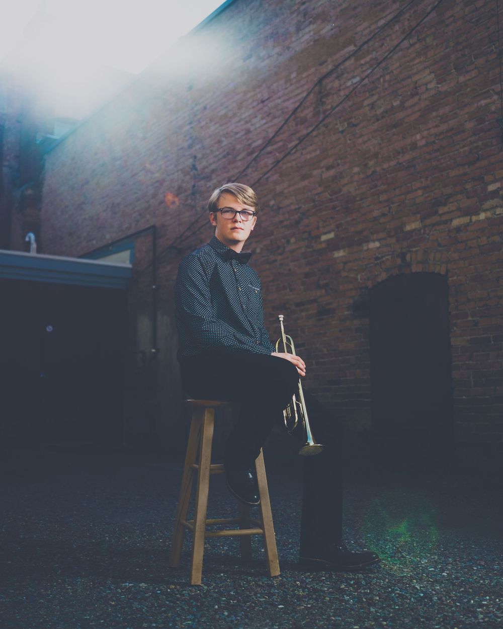 Moody portrait of a musician sitting on a wooden stool against a brick wall holding a trumpet in low light.