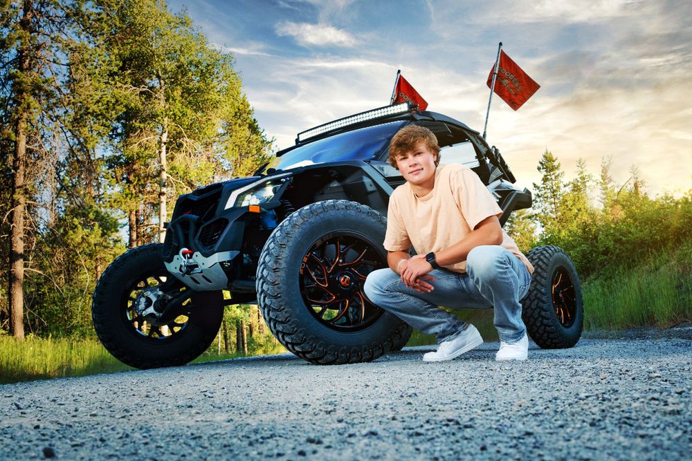 Youth poses next to custom off-road vehicle with red flags on dirt road.