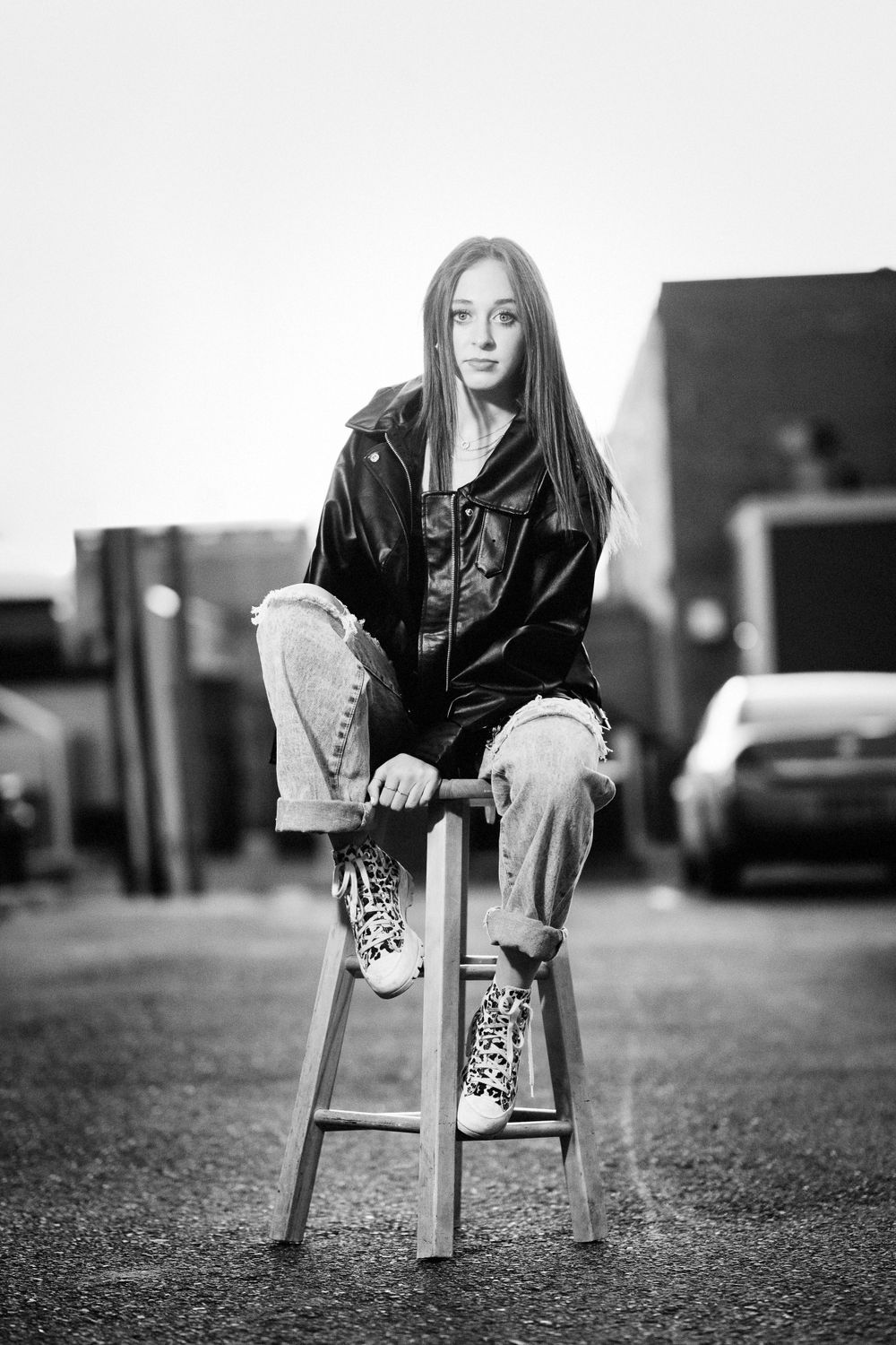 Moody black and white portrait of someone in leather jacket and ripped jeans sitting on wooden stool outdoors.