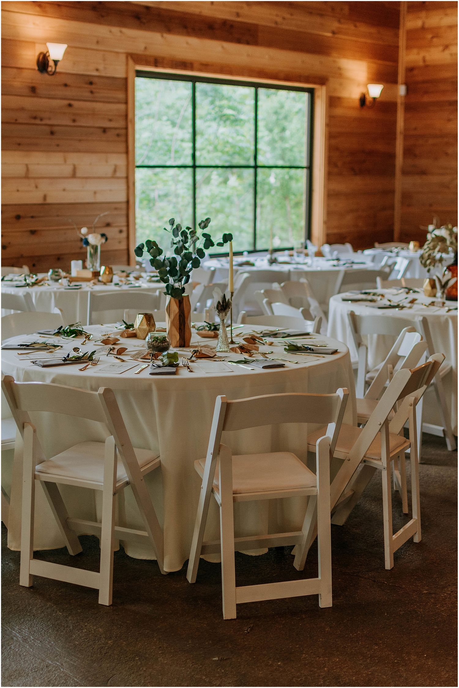 Elegant wedding reception setup with white tables and chairs in a rustic wooden venue with large windows.