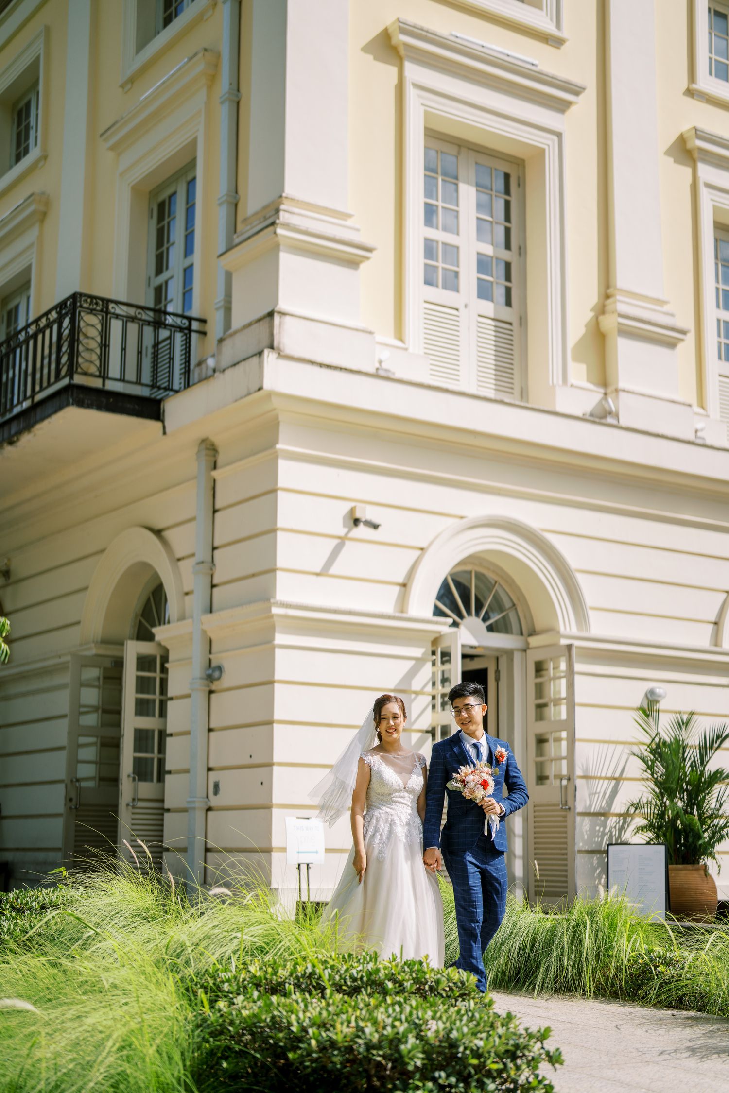 Elegant couple poses outside a grand cream-colored building with classical architecture and arched windows.
