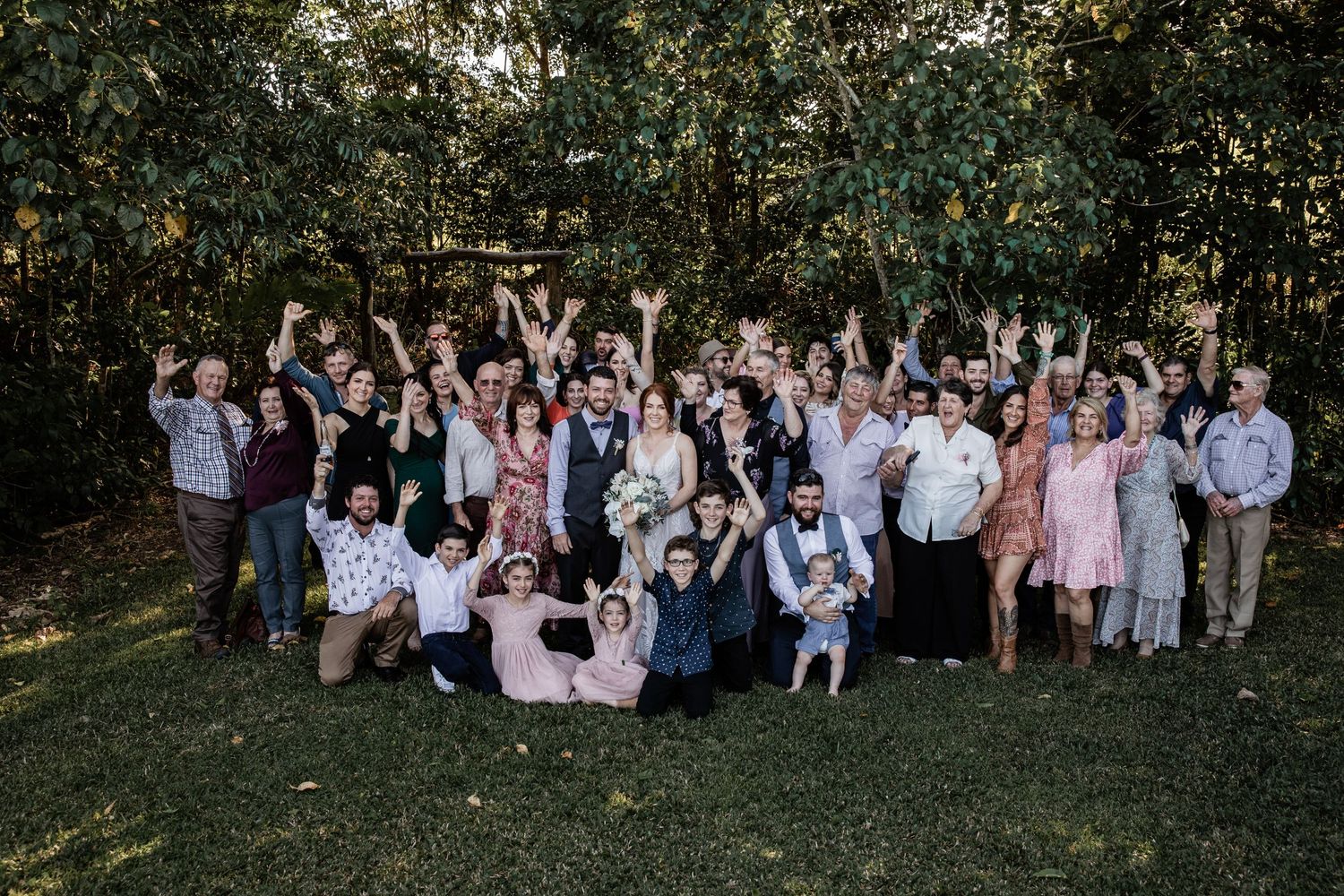A large family gathering poses for a group photo in a wooded outdoor setting.