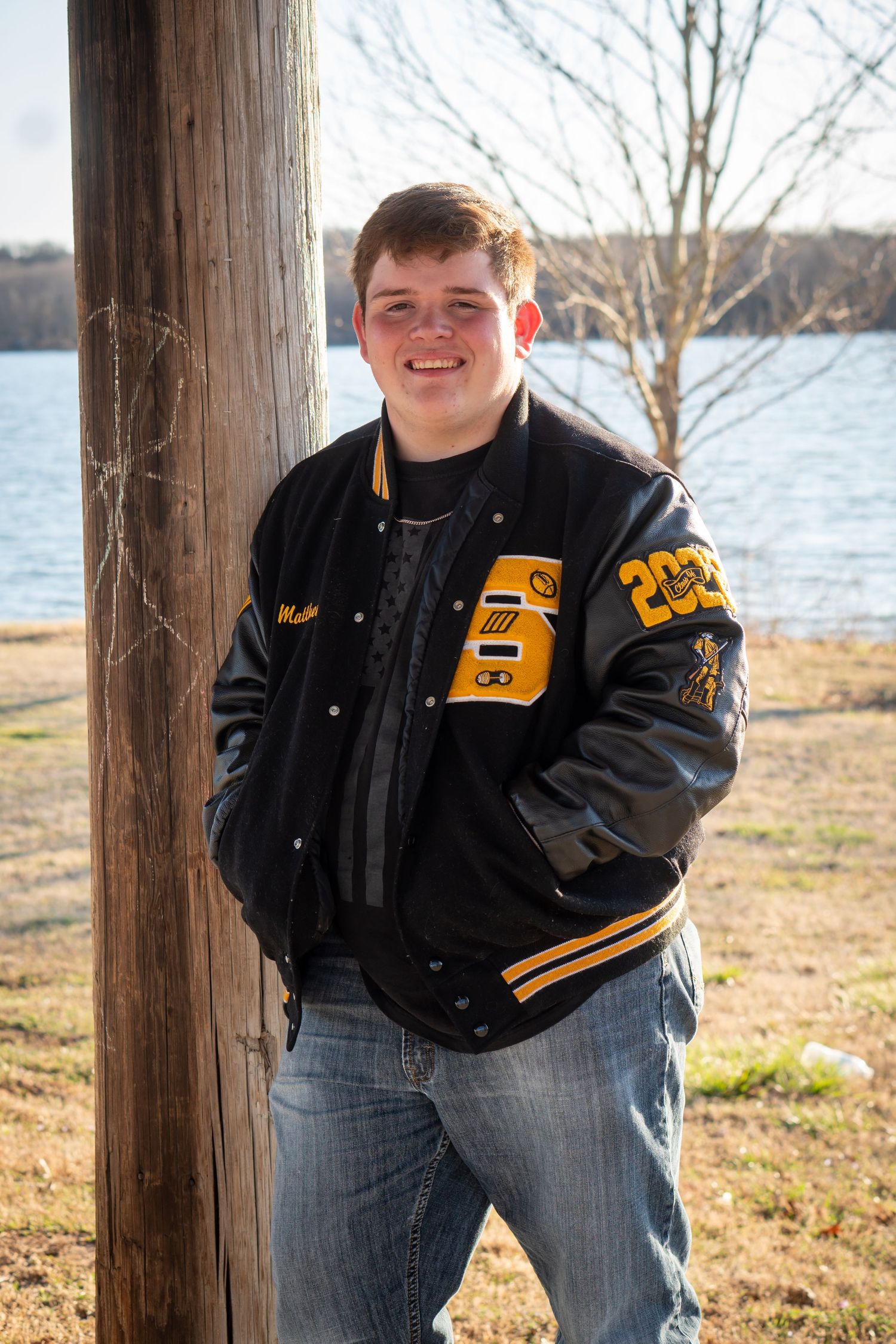 Smiling young man in black and gold varsity letterman jacket leaning against a wooden post near a lake.