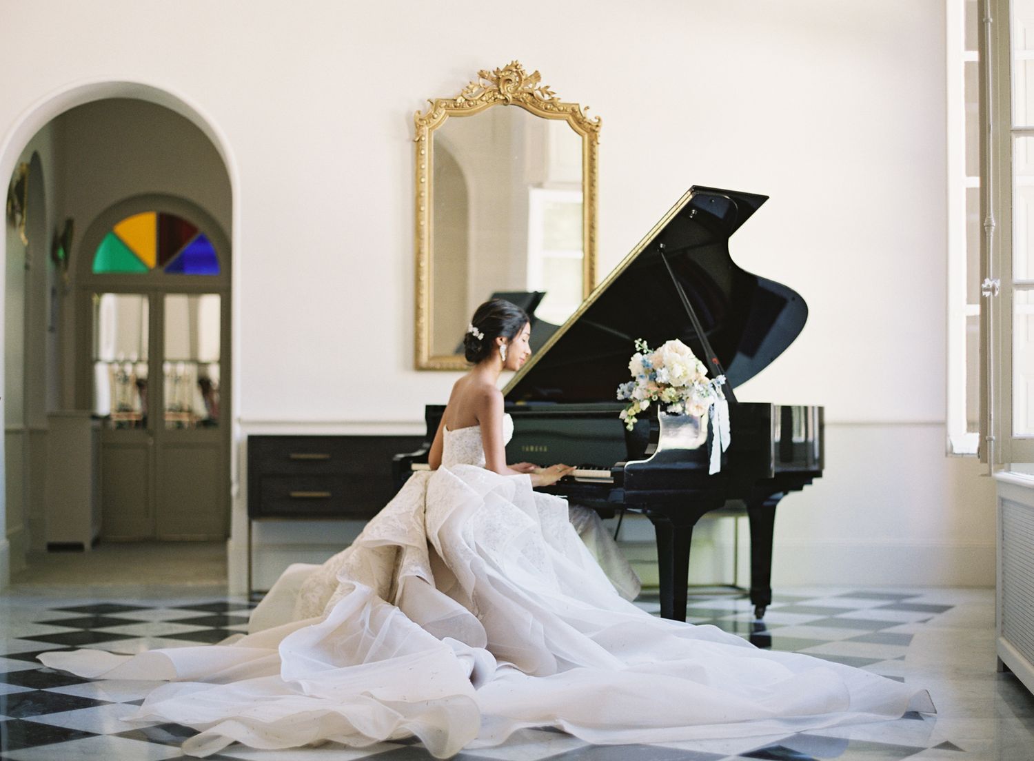 Elegant bridal portrait with flowing white gown at grand piano in classical interior setting.