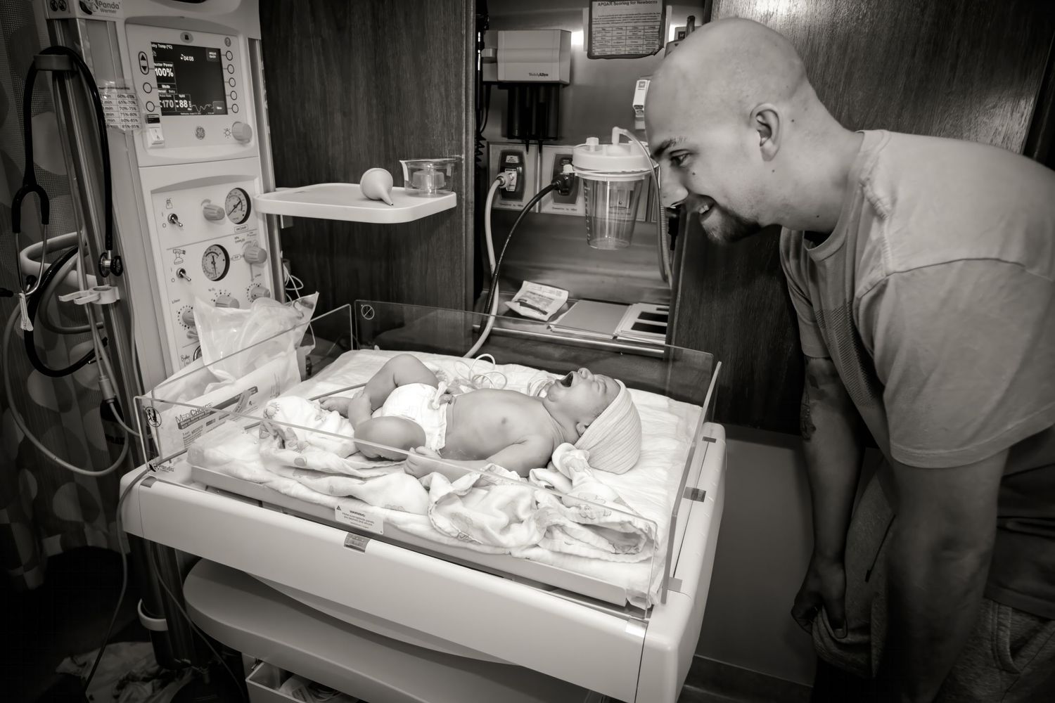 Black and white image of a newborn in a hospital incubator under medical care.