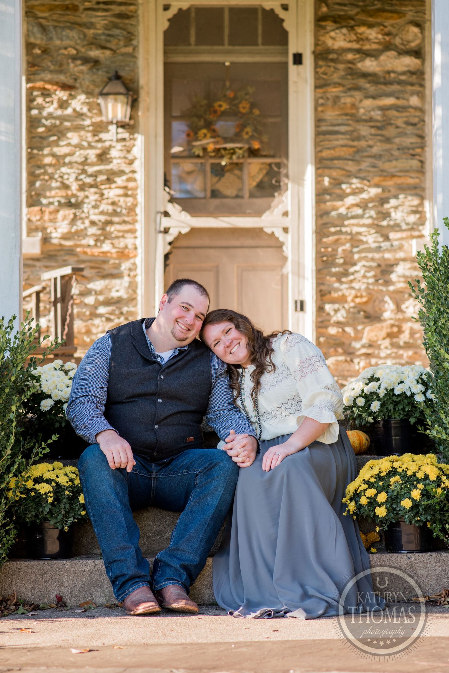 A couple sits on stone steps decorated with yellow mums in front of a rustic stone farmhouse entrance.