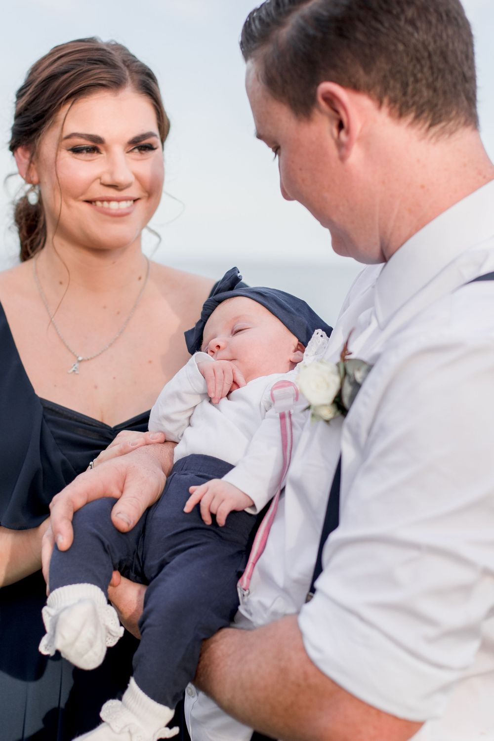 brunette woman in dark blue gown and hair updo smiling at brunette man in suspenders and white shirt holding baby