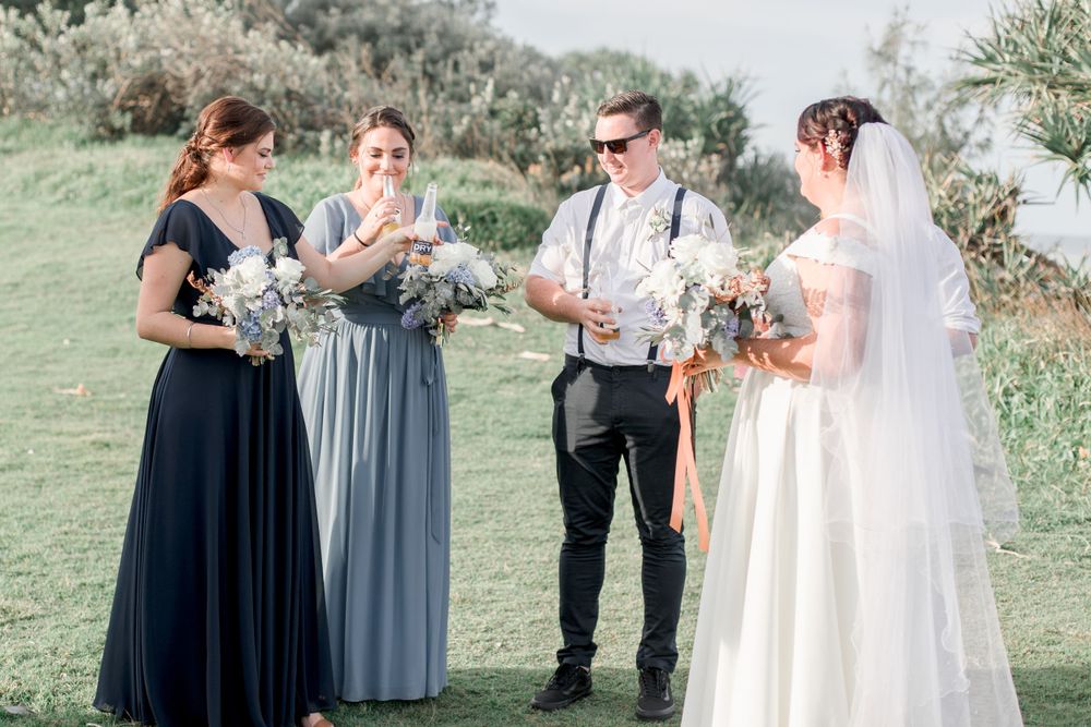 bridesmaids in light and dark blue holding blue and white bouquets drinking champagne with plus size bride on clifftop