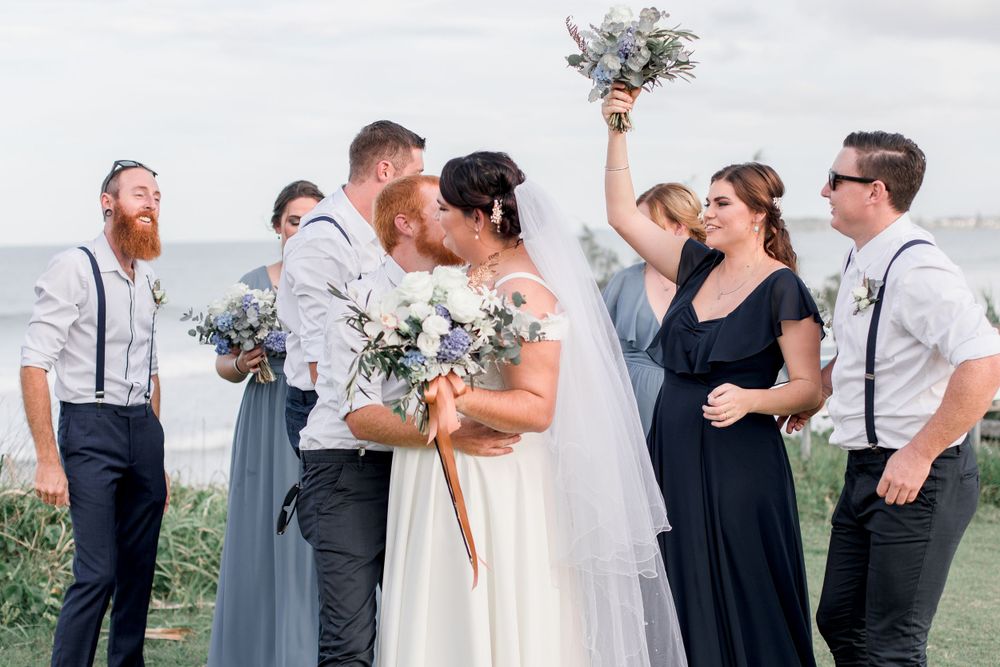 redheaded groom with beard embracing brunette plus size bride with veil and blue and white bouquet ocean in background