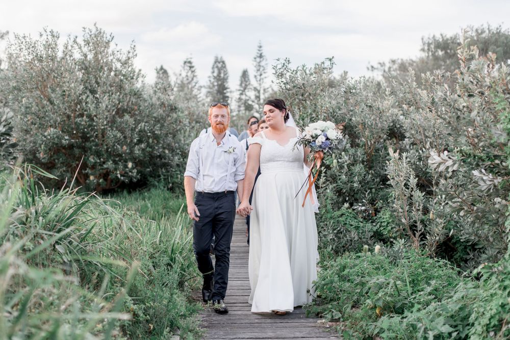 cloudy day with plus size woman in wedding gown and holding hand of groom with beard and casual dress walking on bardwal
