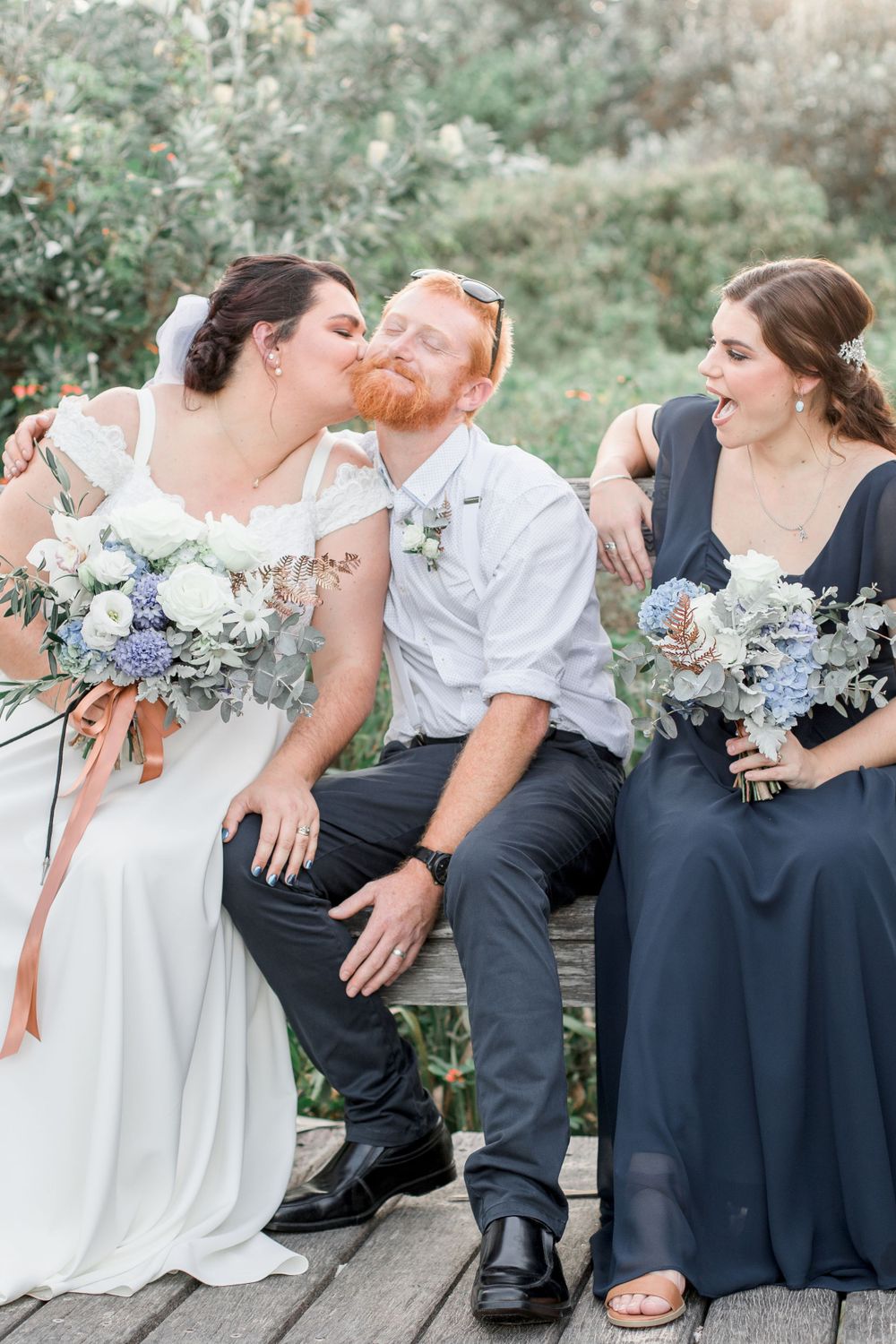 bearded redhead groom in white shirt being kissed by plus size bride while bridesmaid in dark blue laughs