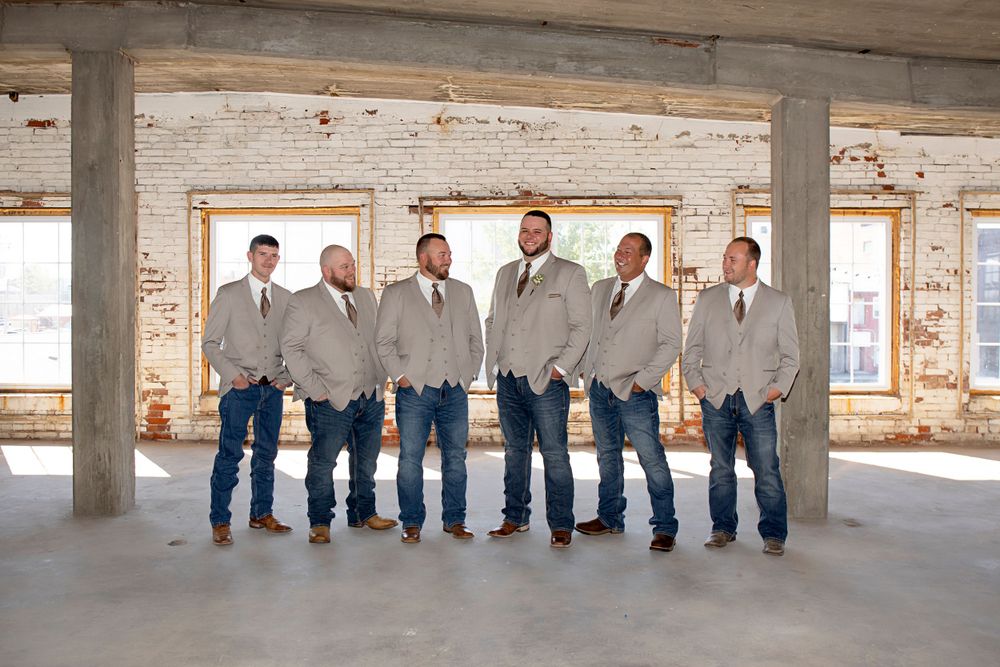 A line of groomsmen in gray suits and jeans pose together in a rustic industrial space with exposed brick walls.