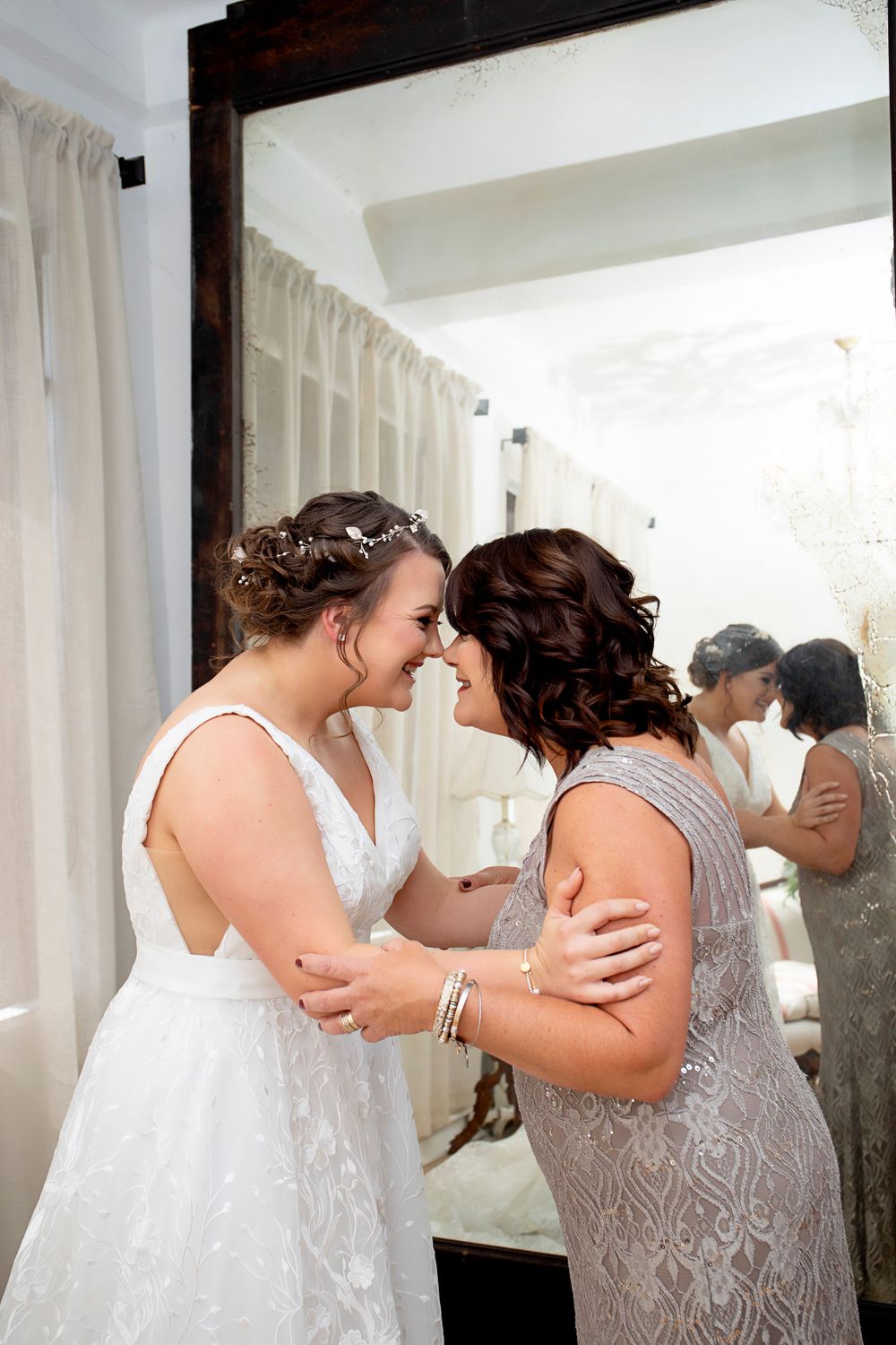Two people share an emotional moment in white lace dresses against a bright interior setting.