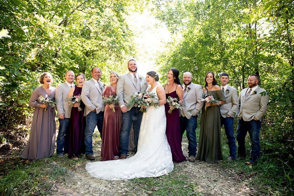 Wedding party poses in shades of gray and burgundy dresses against a lush outdoor forest backdrop.