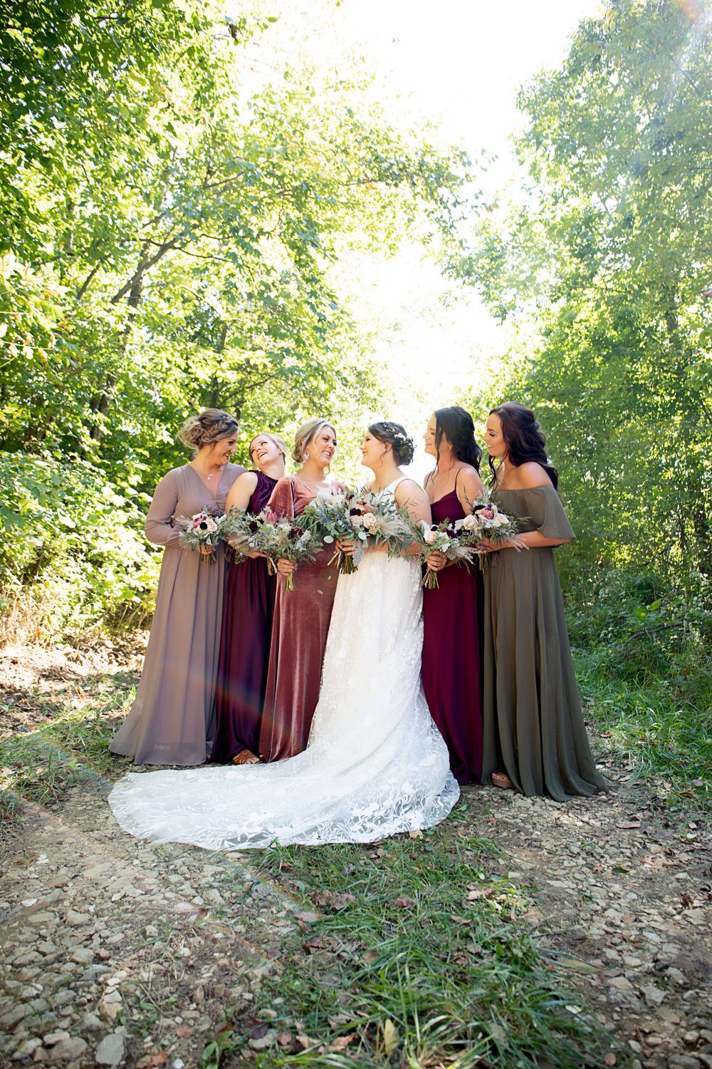 Bridesmaids in flowing burgundy and gray dresses gather around bride in a sunlit outdoor setting.