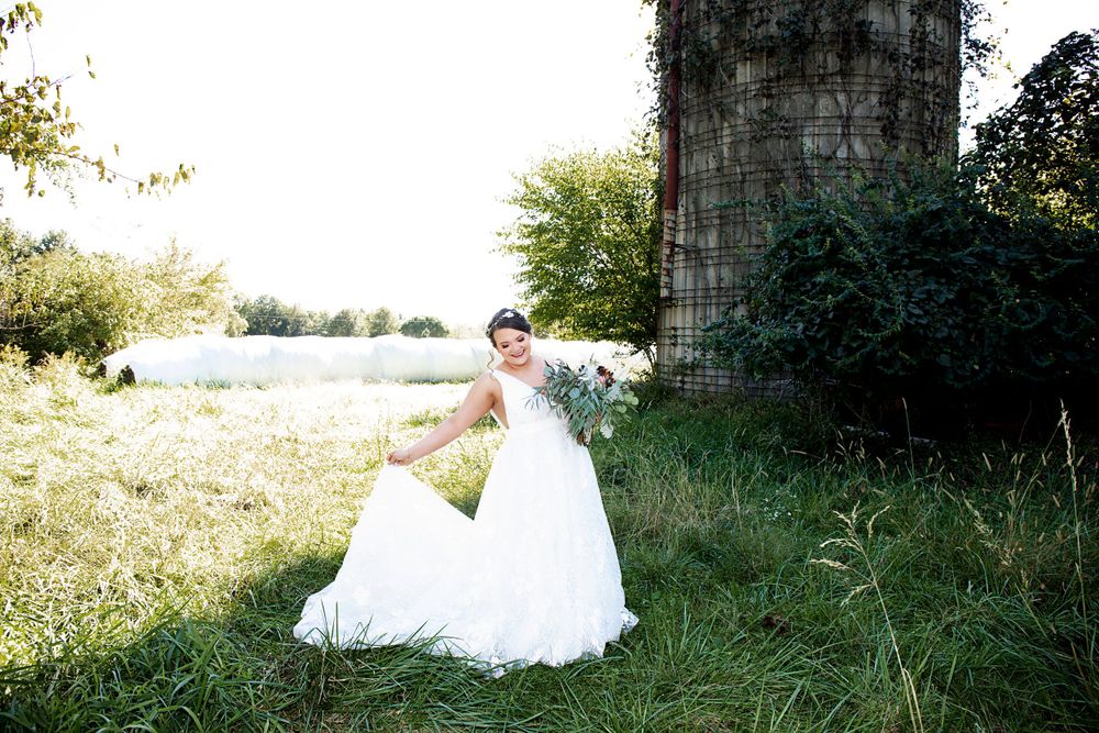 A bride in a flowing white dress poses playfully near a rustic silo structure.