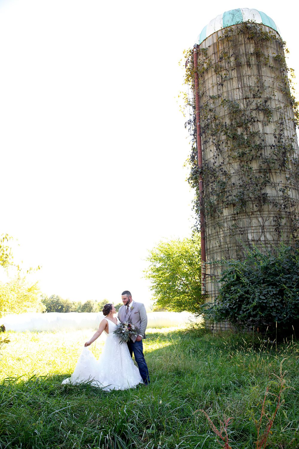 A couple poses romantically by an old silo in a golden-lit field at sunset.