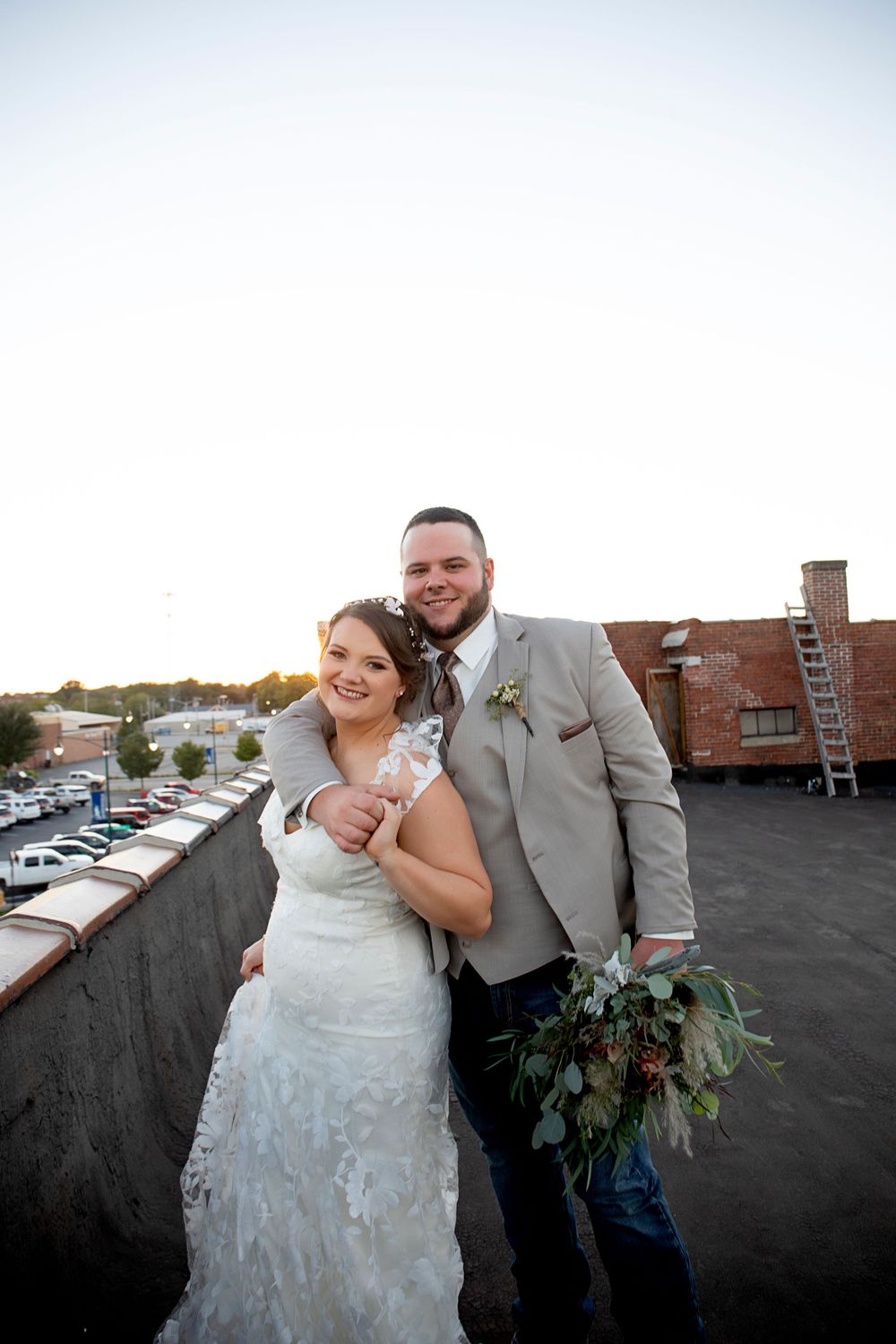 On a rooftop with city views and parked cars below, a newlywed couple shares an intimate moment.
