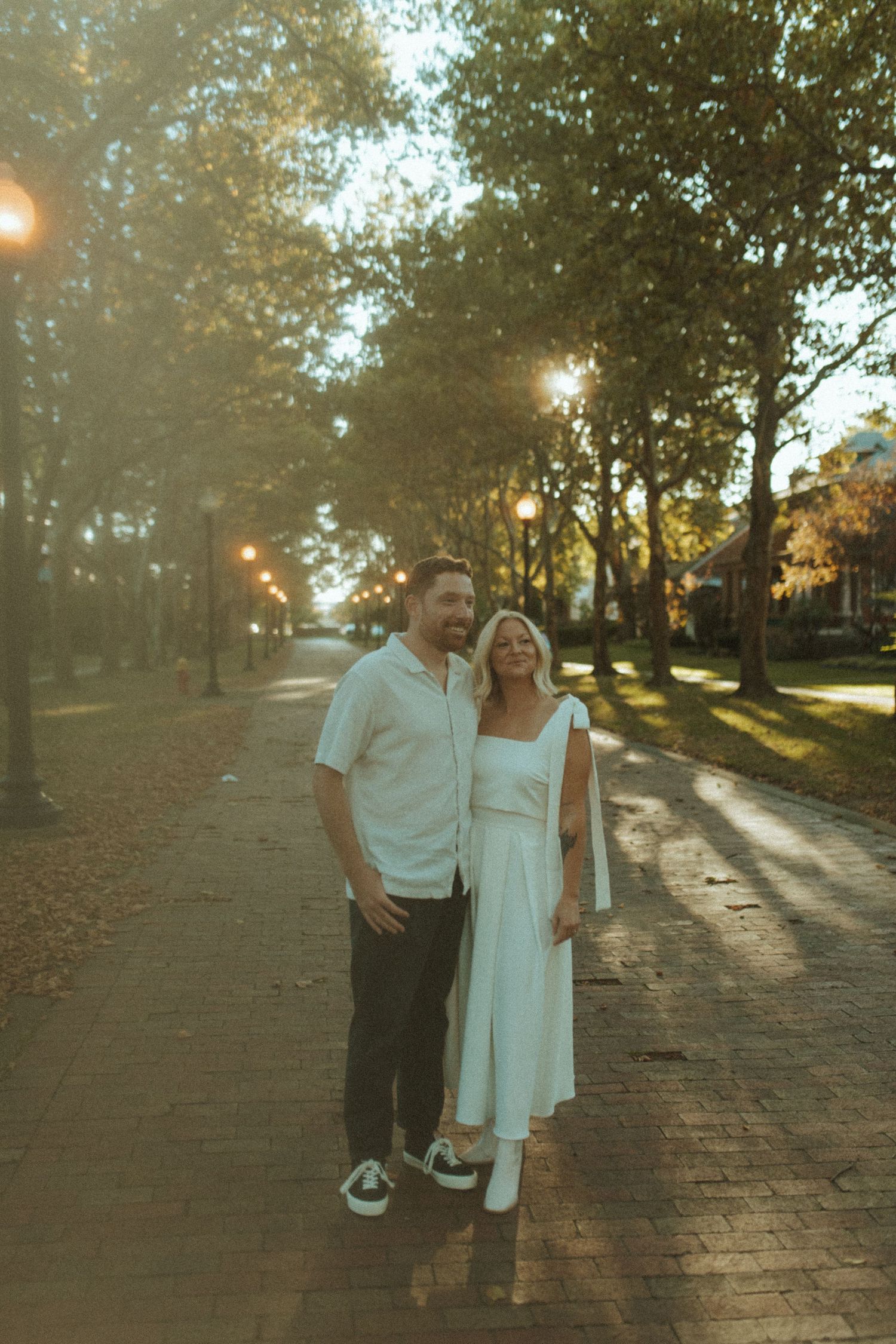 A couple poses on a tree-lined brick pathway at golden hour wearing coordinated white outfits.