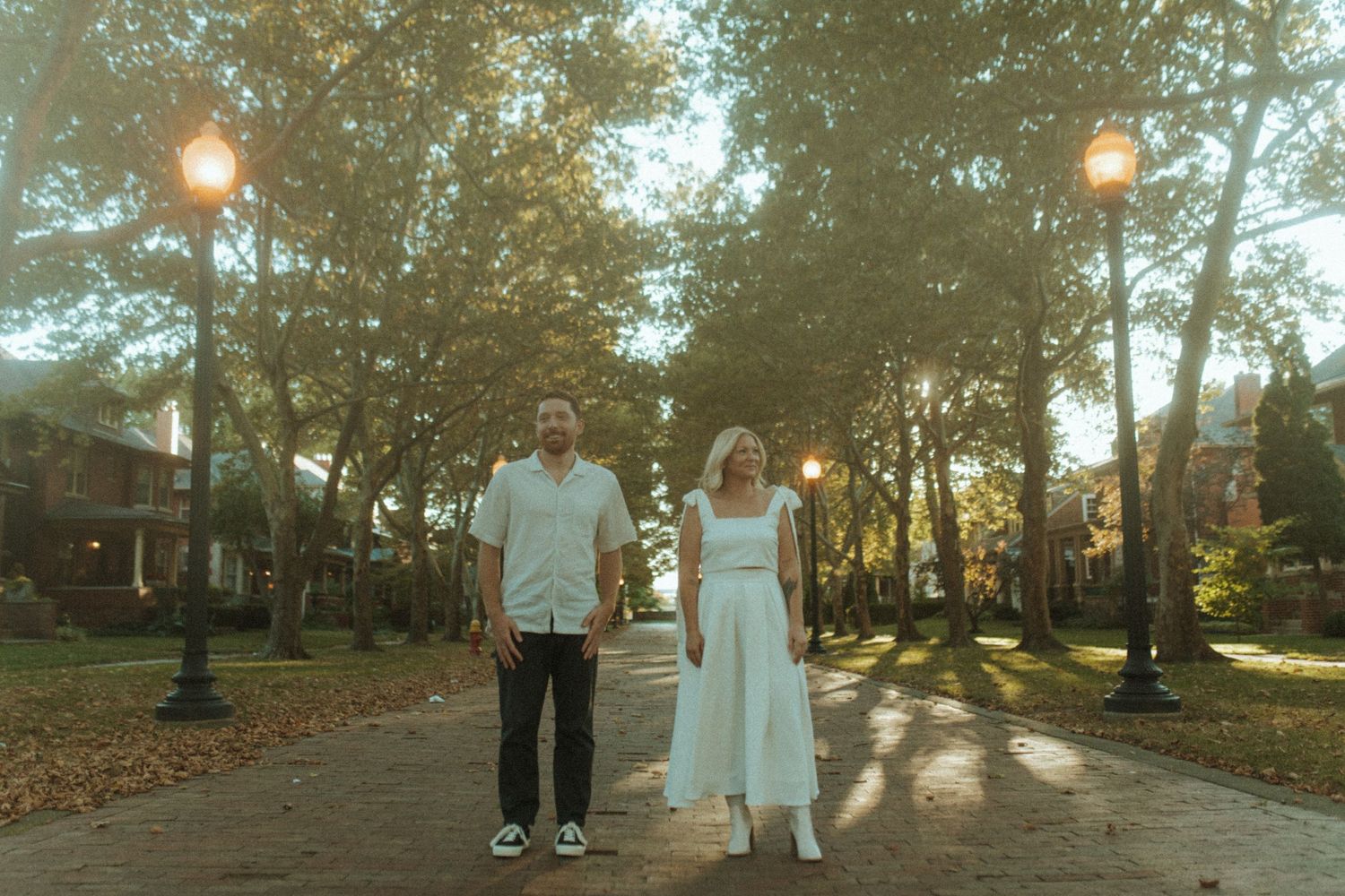Couple in white outfits stands on tree-lined brick pathway with golden sunlight filtering through.