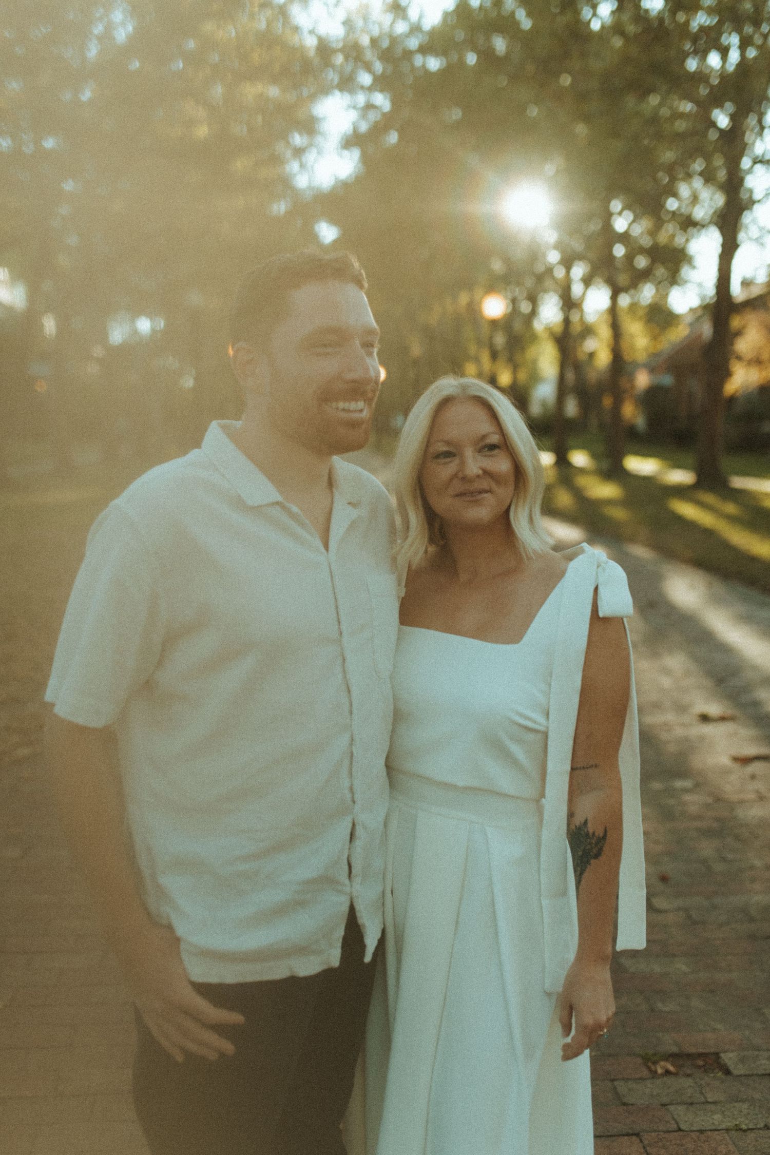 Smiling couple stands close together on brick walkway with sunlit trees in background.