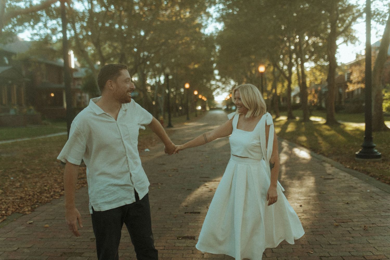 Couple dances playfully on brick pathway surrounded by autumn trees and streetlights.