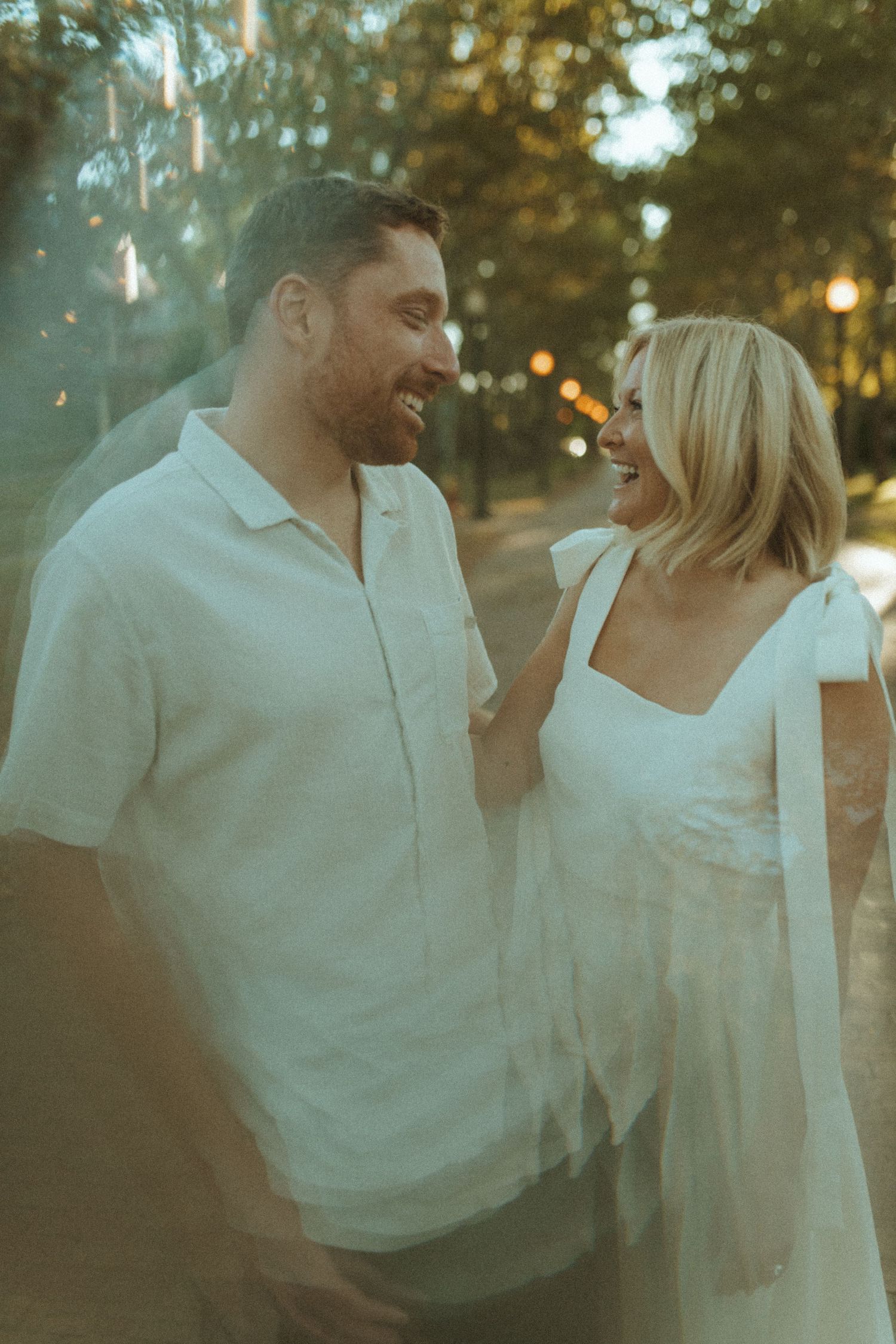 A romantic couple in white outfits shares a tender moment at sunset in a park setting.