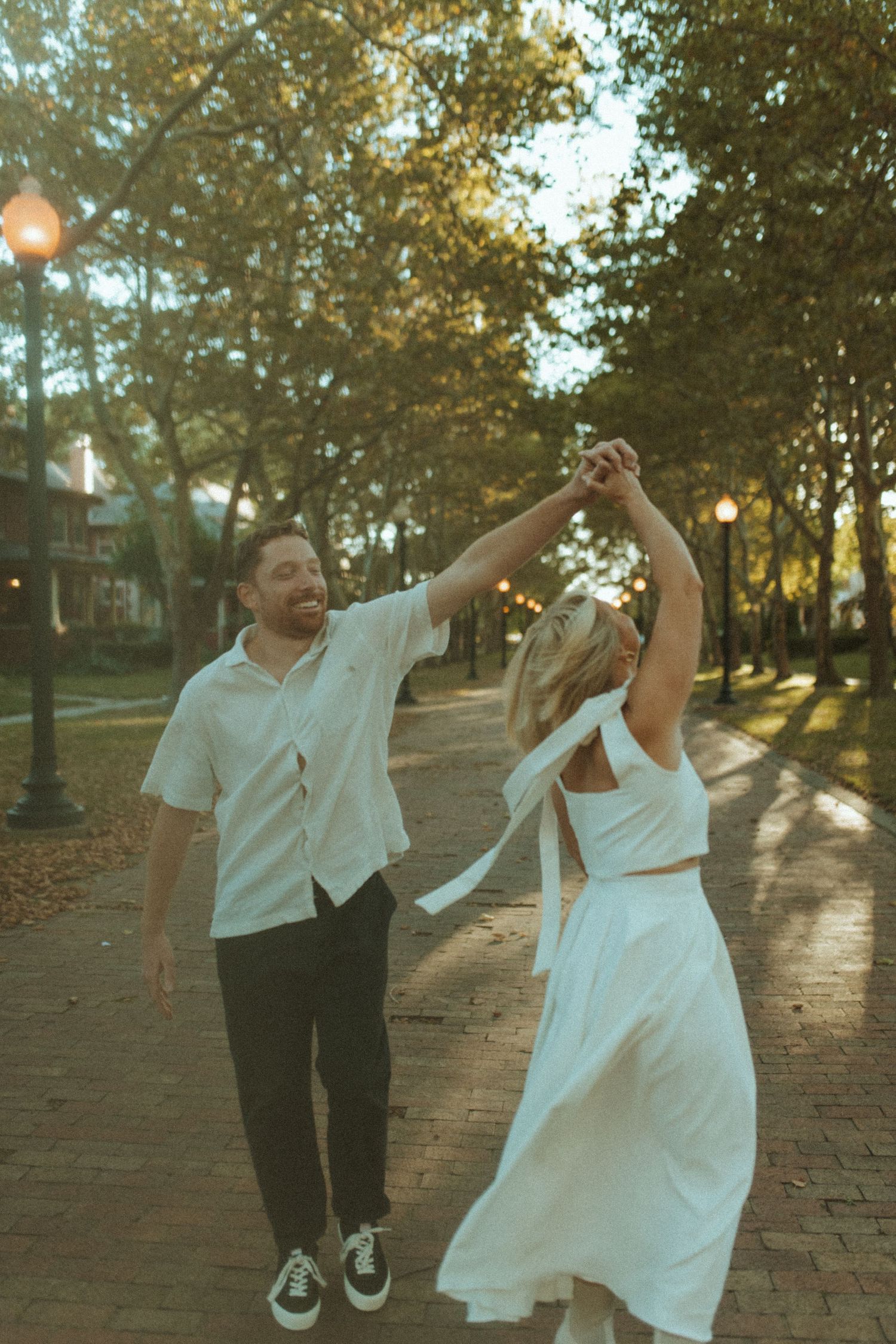 Dancing couple in white outfits twirls on brick pathway during golden hour.