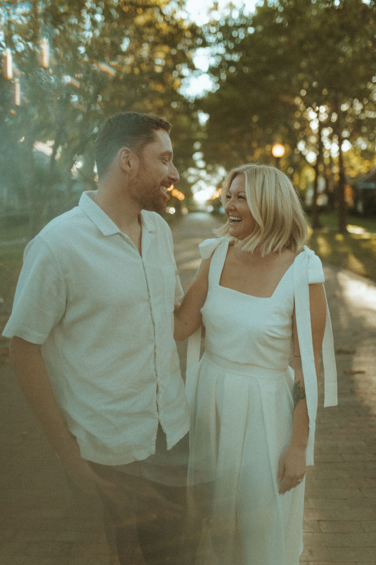 Couple shares intimate laugh while strolling on tree-lined pathway at sunset.
