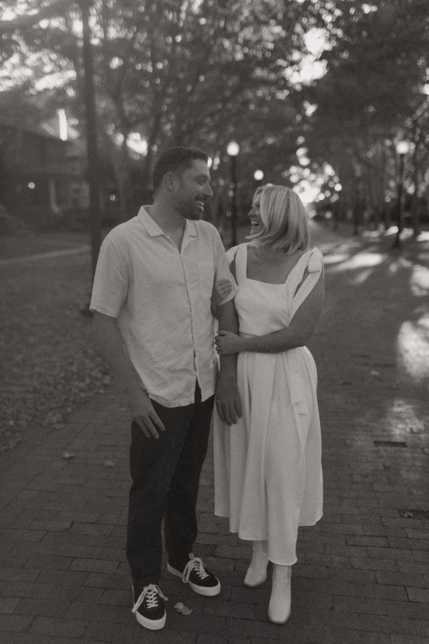 Black and white photo of couple in coordinated outfits standing on brick path.