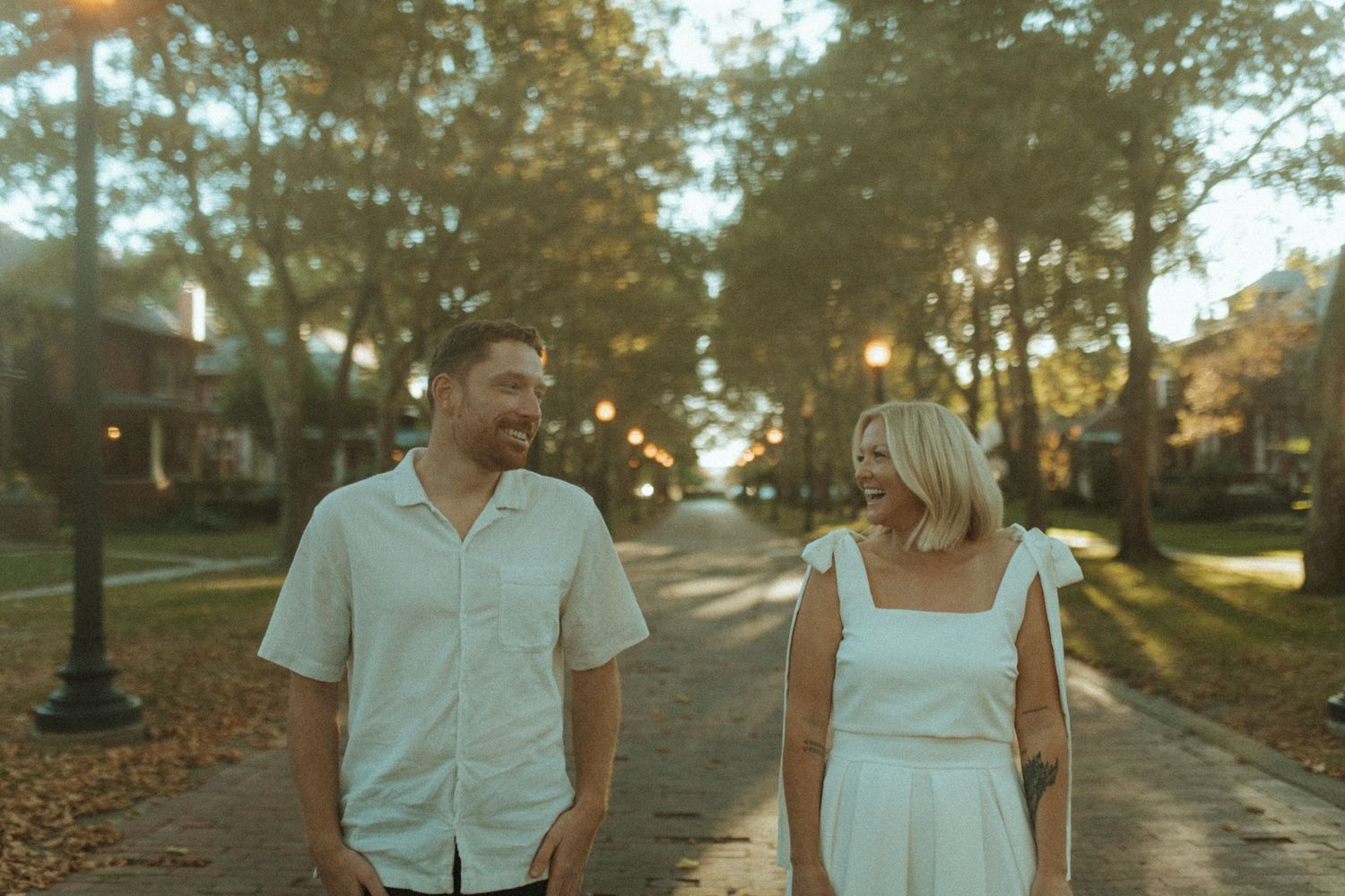 Couple shares candid moment while walking along brick pathway with sun-dappled trees.