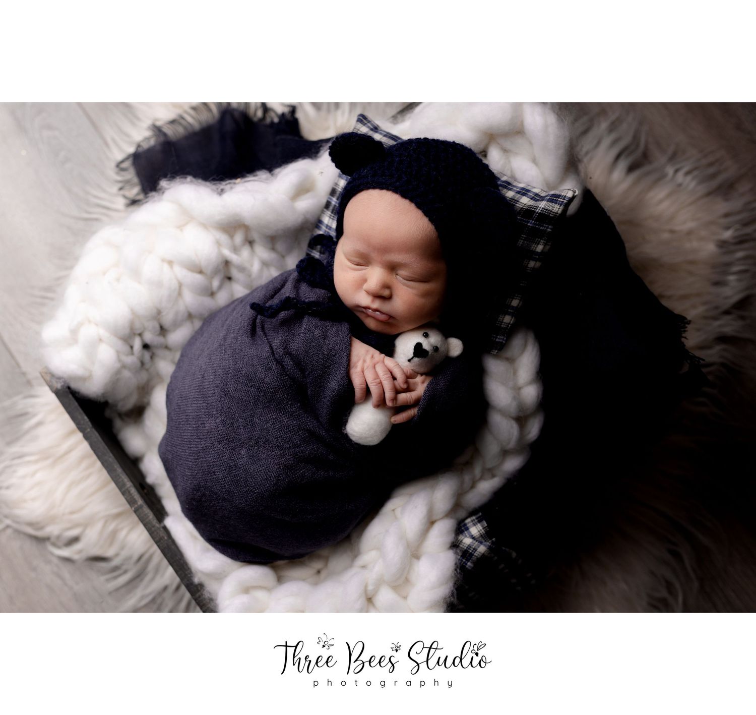 Newborn in navy outfit and bear hat sleeping on white fur background.