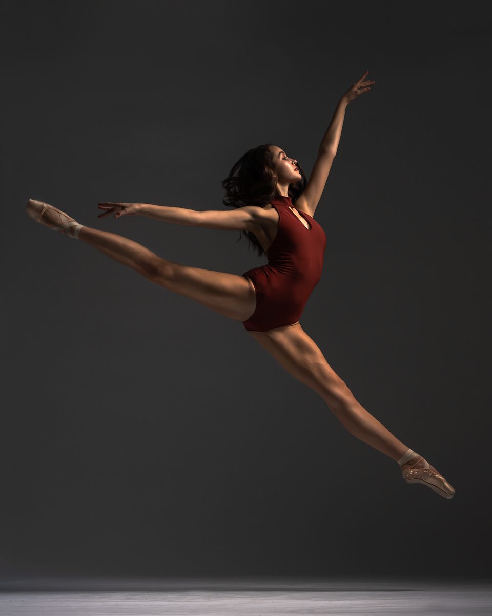 Dynamic ballet leap captured mid-air with dancer in burgundy leotard against dark backdrop.