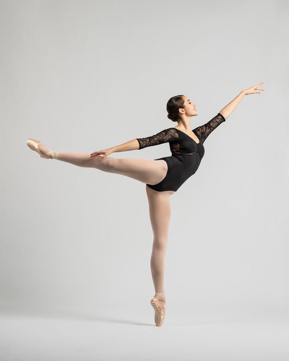 Ballet dancer executes graceful arabesque in black lace leotard against light background.