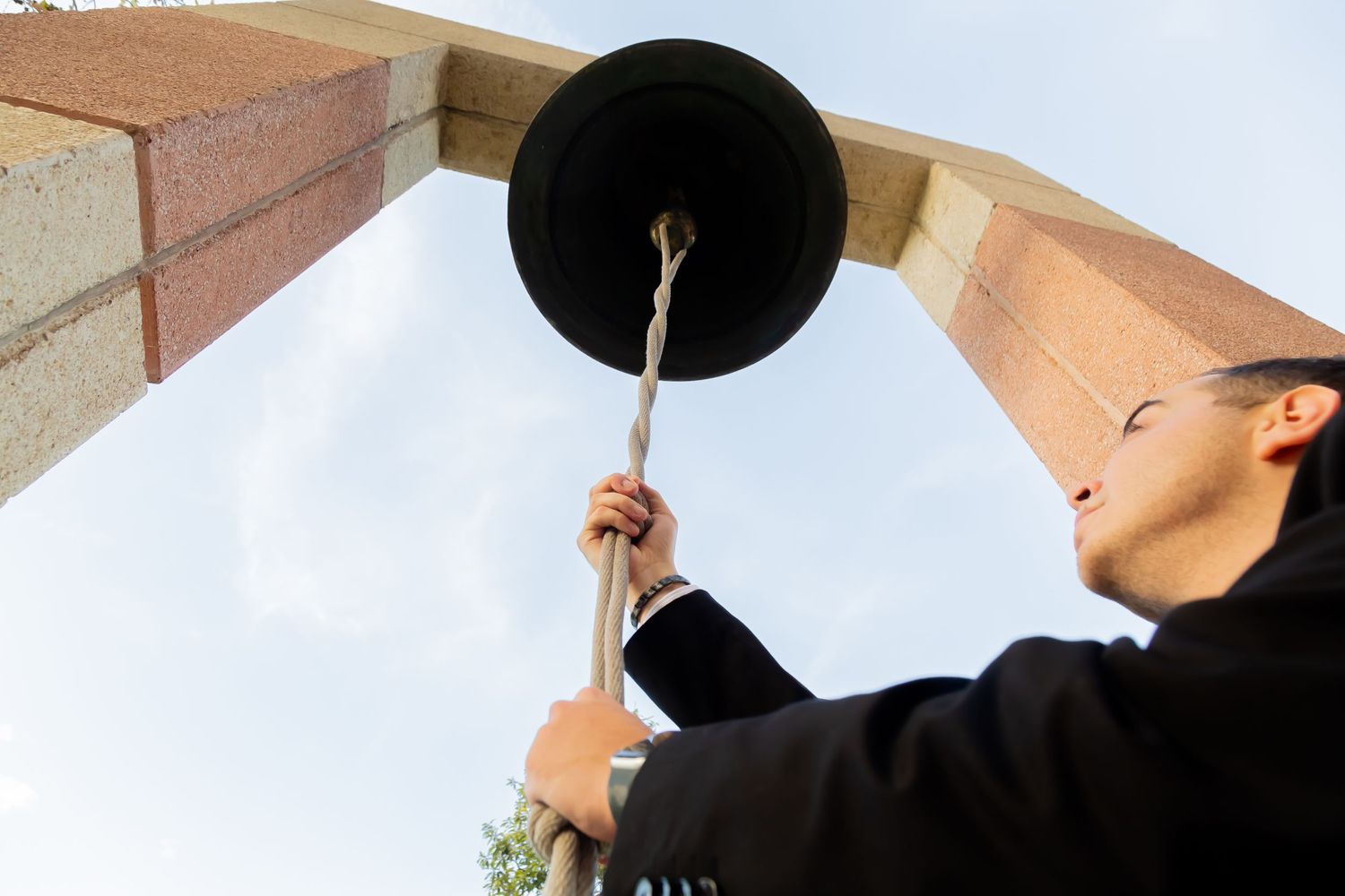 Young man in black suit rings a large bell between brick columns against a blue sky.