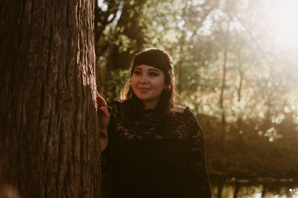 Person in black winter attire stands beside large tree trunk in golden forest light.