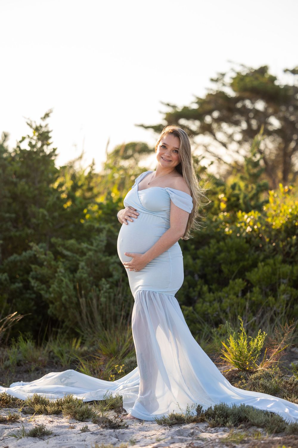 An expectant mother in a flowing white gown poses at sunset among coastal vegetation.