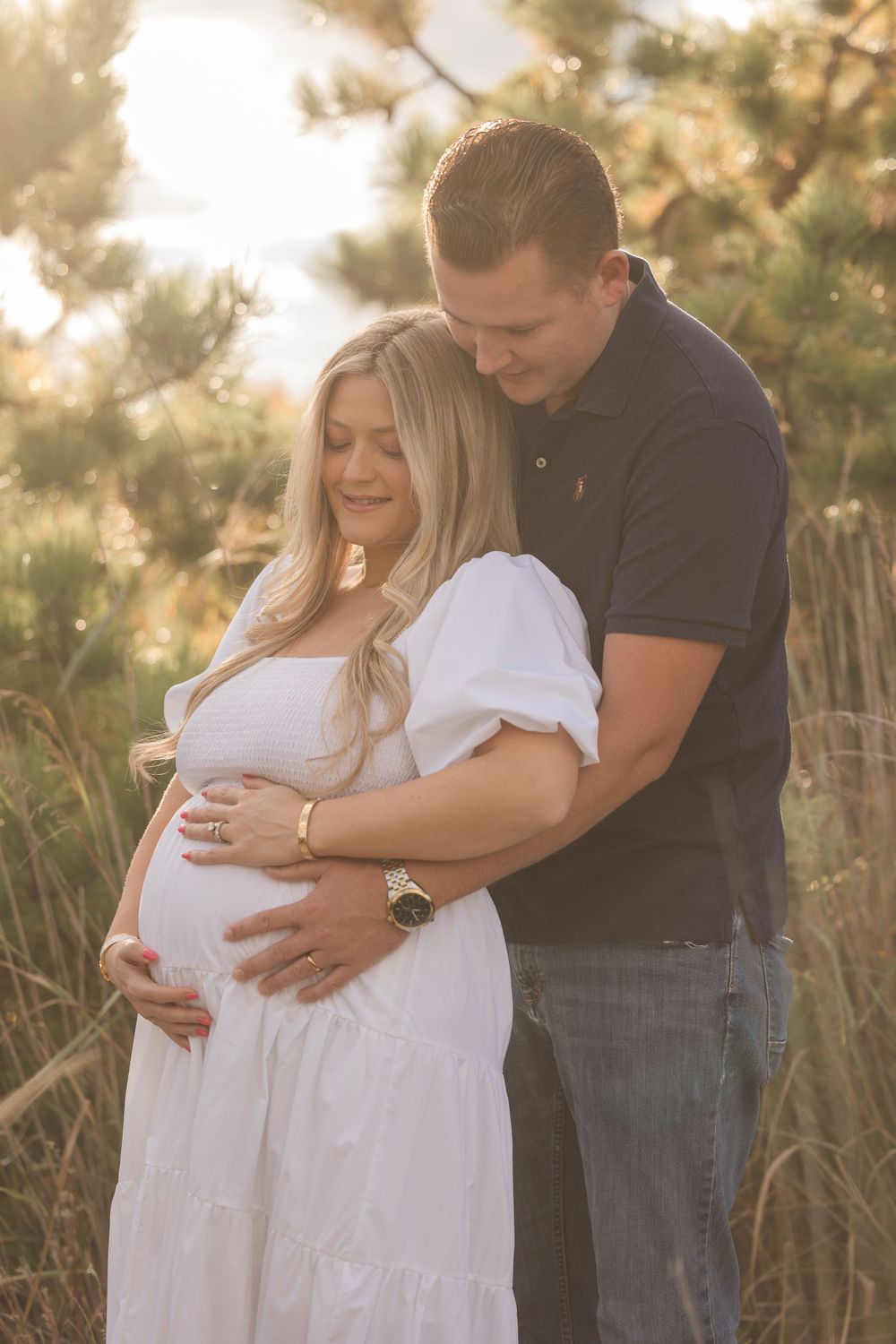 Golden hour maternity photo with couple embracing among tall grass.