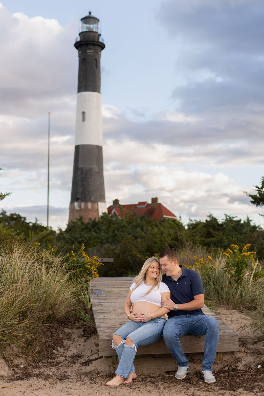Maternity photos taken near a lighthouse with coastal dunes and beach grass in the background.