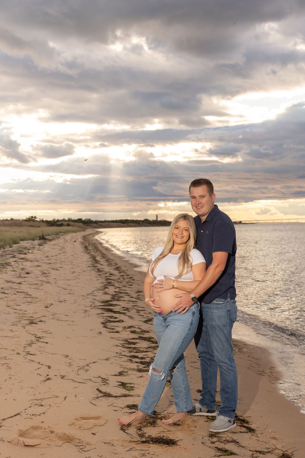 Maternity photoshoot on a sandy beach at sunset with dramatic clouds and ocean views.
