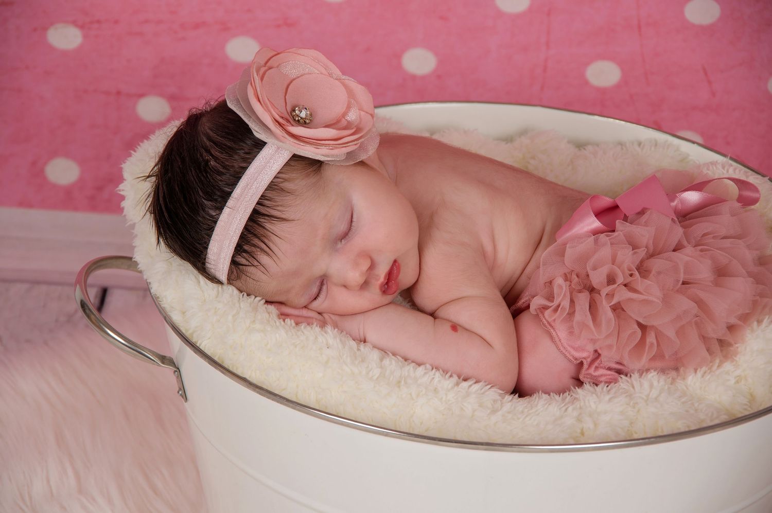 newborn photography of baby sleeping in a bucket with pink backdrop and wearing pink bloomers and headband