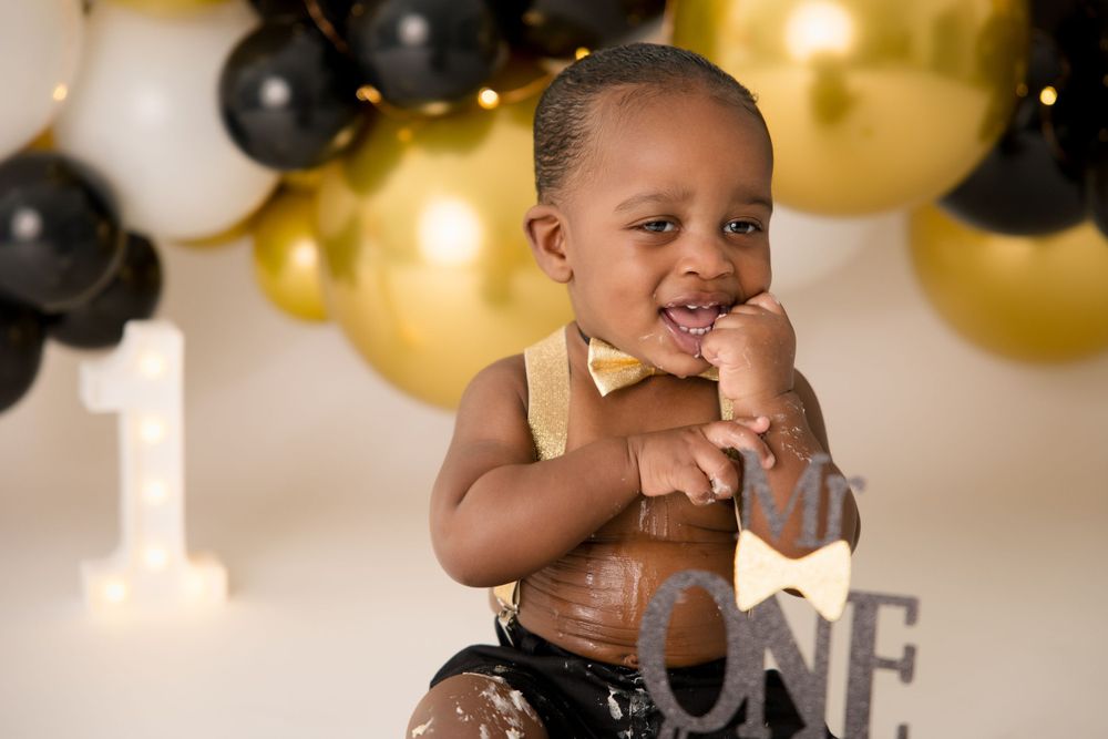 Baby in a gold outfit poses against a backdrop of black and gold balloons for a first birthday shoot.