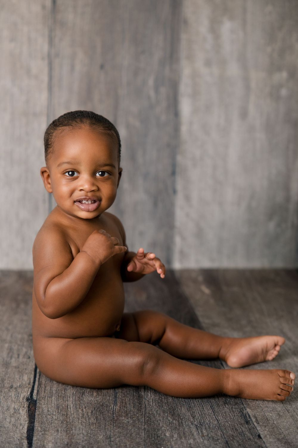 Baby sitting on wooden floor during minimalist photography session against grey backdrop.