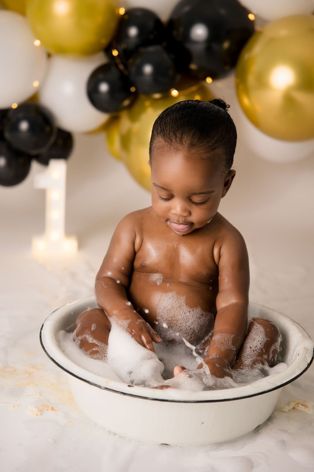 Baby sits in soapy water during a bath photoshoot with festive balloons in the background.