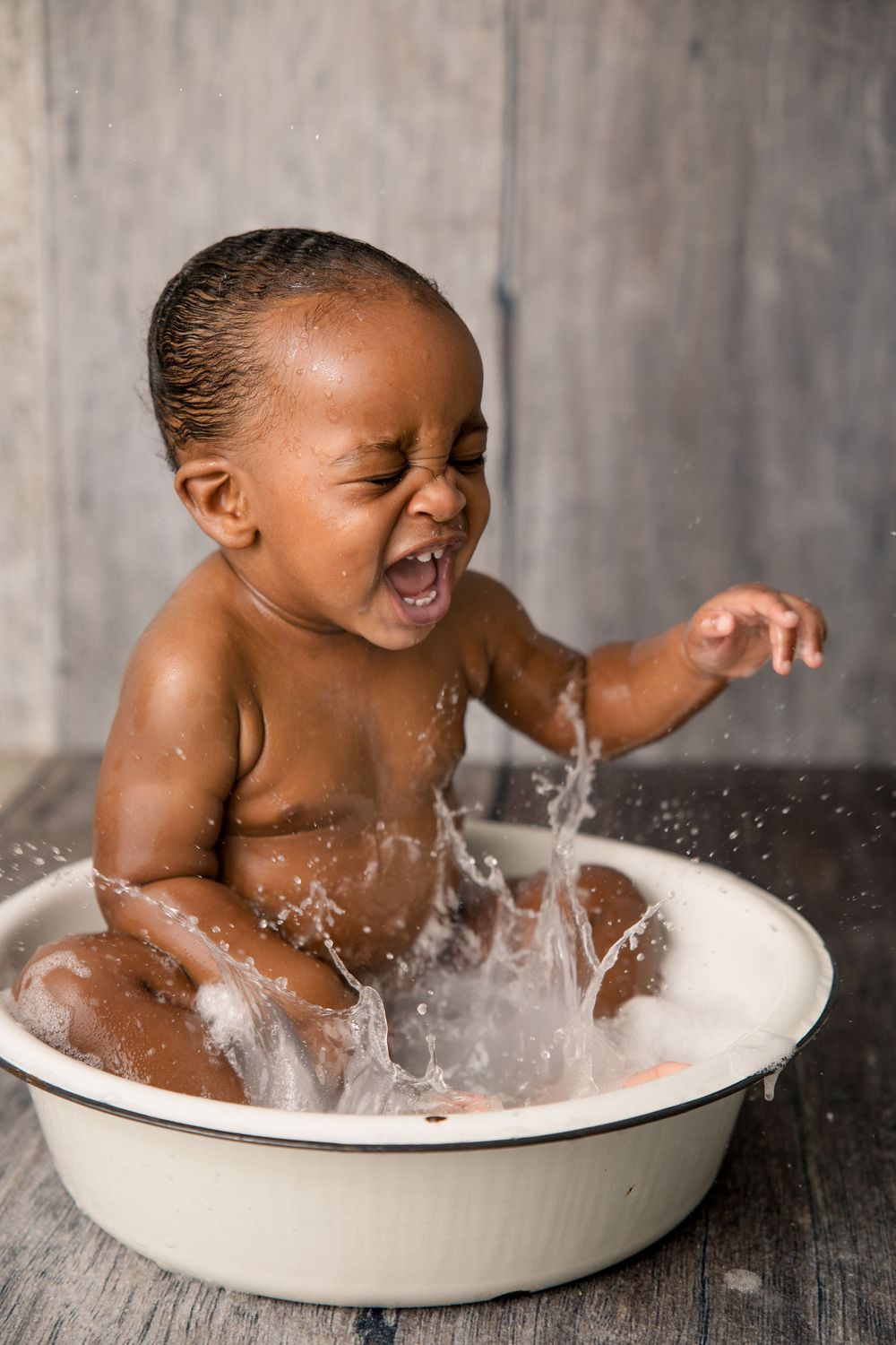 Baby laughs while splashing water during a bath in a white basin against a grey backdrop.