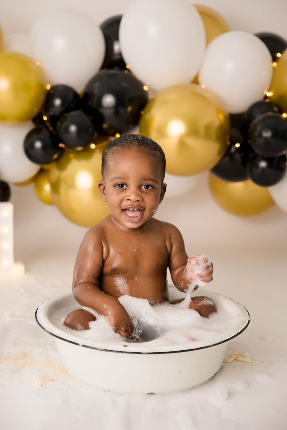 Happy baby splashes in a white basin surrounded by gold and black balloons during a celebration.