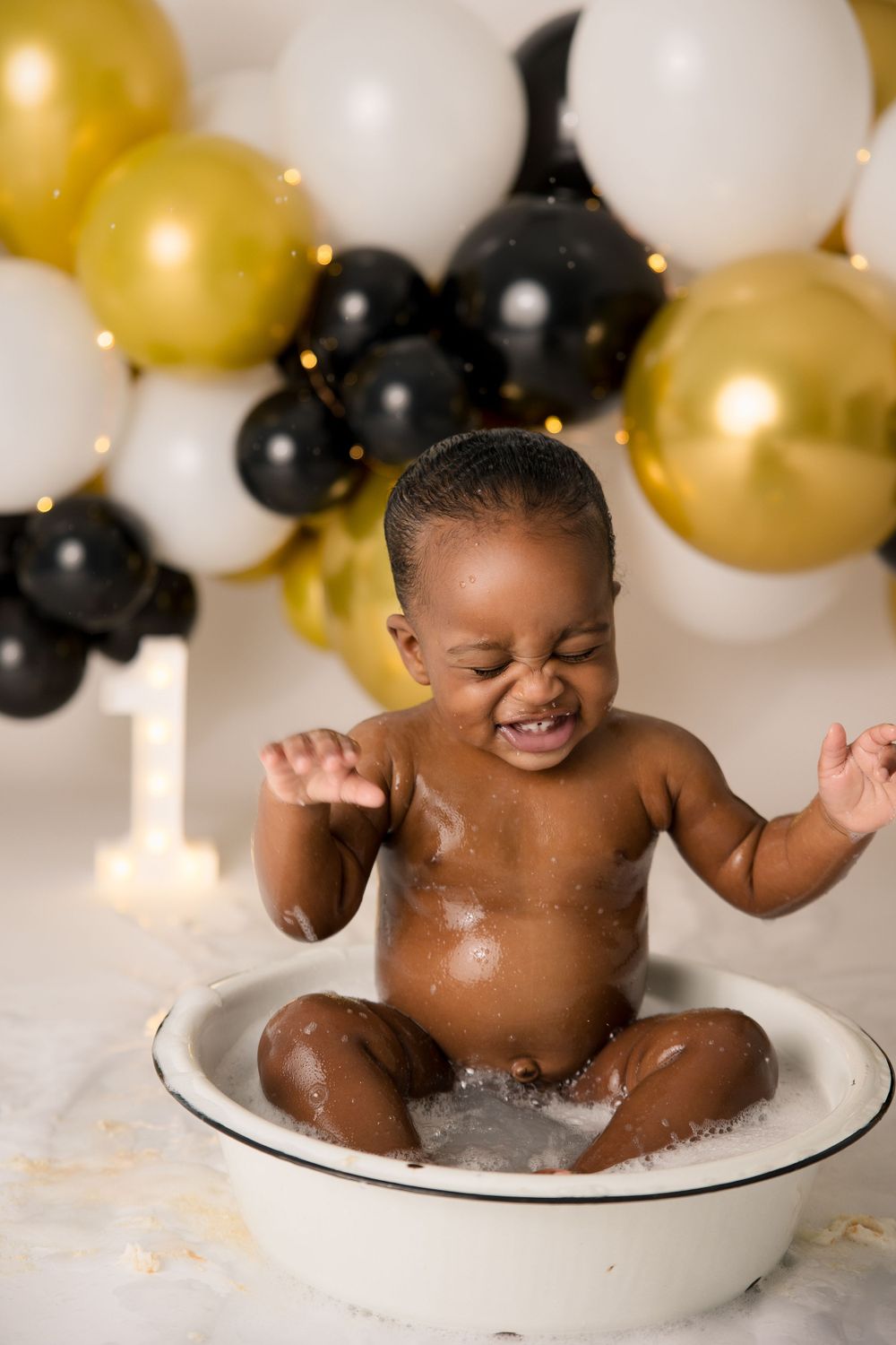 Joyful baby splashes in a white basin during a celebratory photoshoot with balloons.