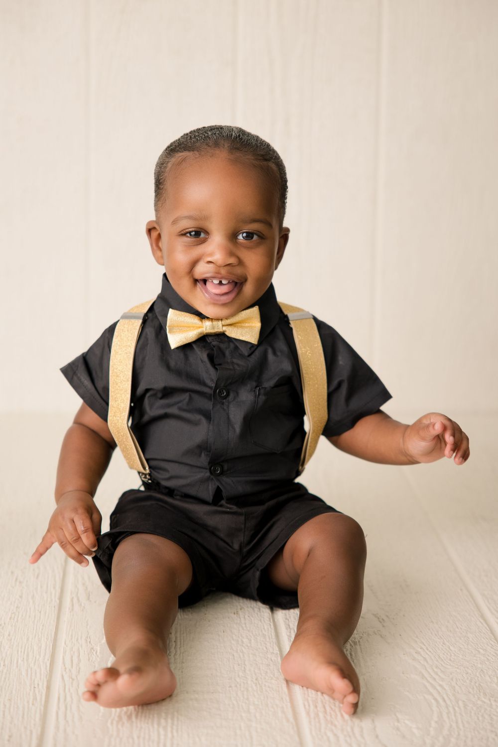 Baby in black outfit with gold bow tie sitting on white floor during portrait session.