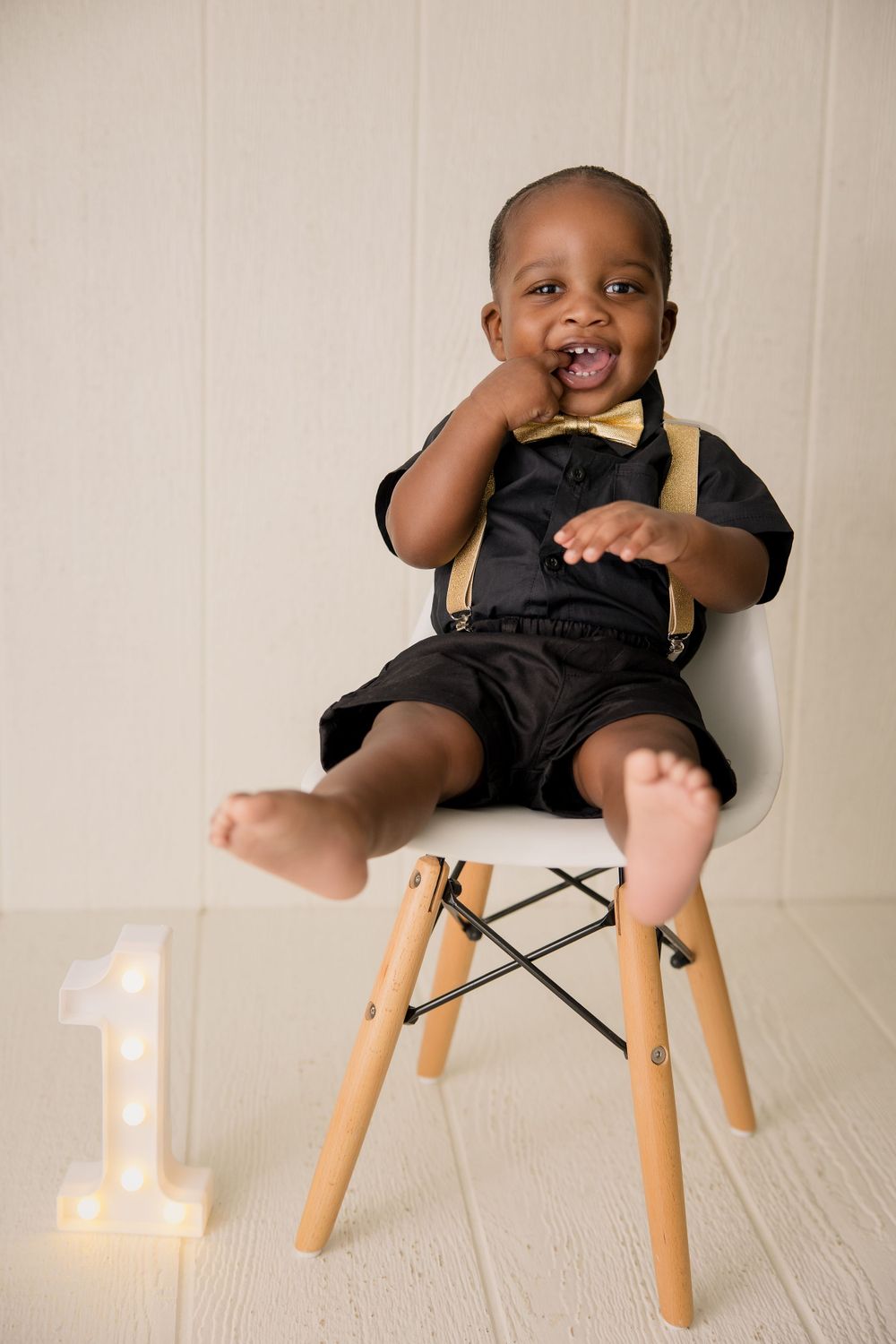 Baby in formal attire sitting on wooden chair next to illuminated number one.