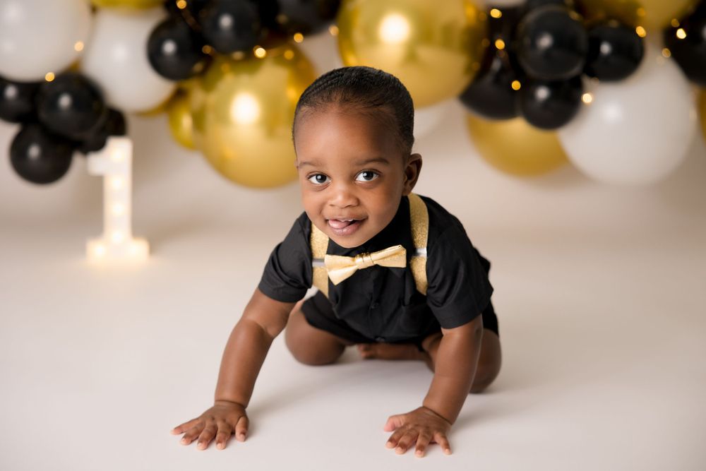 Baby in formal wear crawling under gold and black balloon arch decoration.